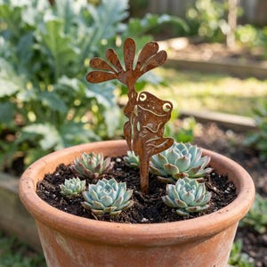 Puede incluir: Un estaca de jardín de metal color óxido con forma de rana sosteniendo una flor, colocada en una maceta de terracota con tierra oscura y cinco plantas suculentas de color verde claro. El recorte de metal presenta una silueta de rana estilizada.