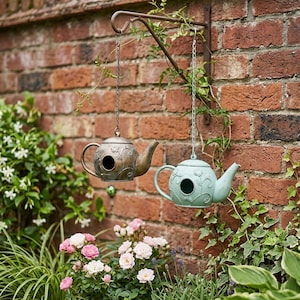 Könnte beinhalten: Zwei teekannenförmige Vogelhäuser hängen an einer Wandhalterung aus Metall. Ein Vogelhaus ist metallisch bronzefarben mit geprägten Blumenmustern, das andere ist hellblau mit ähnlichen Blumendetails. Beide haben ein kreisförmiges Eingangsloch.