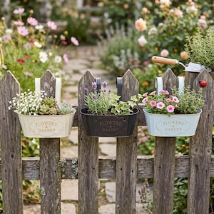 Peut inclure: Trois jardinières suspendues ovales en métal de couleur crème, noire et bleu clair, avec un support blanc et l'inscription en relief "FLOWERS & GARDEN". Elles contiennent diverses plantes vertes, du gypsophile blanc et des fleurs roses.