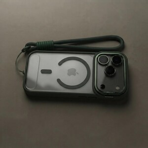 May include: A dark green smartphone case with a clear back panel featuring a circular magnetic ring and a rectangular camera cutout. The case includes a matching dark green woven wrist strap attached to the top corner for secure carrying.