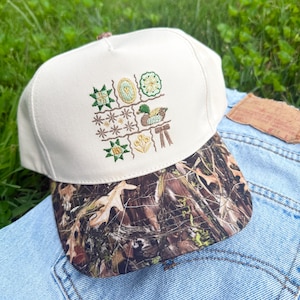 May include: A cream-colored baseball cap with a camouflage-patterned brim. The front panel features an embroidered grid design containing various small motifs, including green stars, a yellow oval, a mallard duck, and a small yellow flower.