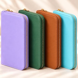 May include: Four rectangular zippered cases standing upright in a row. The cases are colored lavender, dark green, brown, and light blue. Each case features a smooth exterior surface, rounded corners, and a gold-toned metal zipper closure.