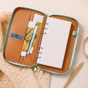 May include: A light blue zippered planner with a tan interior featuring multiple card slots, document pockets, and a six-ring metal binder mechanism holding blank white paper. A gold pen rests on the side, and a blue card is tucked into a slot.