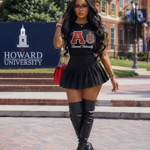 Puede incluir: Una camiseta negra de manga corta con una letra A roja y el texto Howard University, combinada con una minifalda plisada negra y botas negras por encima de la rodilla. Un bolso rojo con correa de cadena dorada se lleva al hombro.