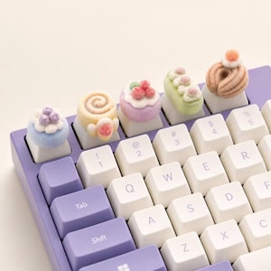 Puede incluir: Un teclado mecánico morado con cinco teclas artesanales personalizadas en forma de postres en miniatura, incluyendo un pastel azul, un rollo beige, una tarta de bayas morada, una barra glaseada verde y una dona beige con un pequeño adorno rosa.