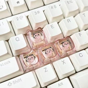 May include: Three square, translucent pink mechanical keyboard keycaps featuring a cartoon cat face graphic. Each keycap displays a different facial expression for the cat, including a winking eye, a smiling mouth, and a neutral look, set against a white keyboard.