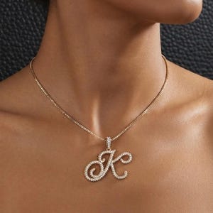 May include: A gold-toned chain necklace featuring a cursive letter K pendant. The pendant is encrusted with numerous small, clear, sparkling stones that cover the entire surface of the letter, creating a bright and reflective appearance.