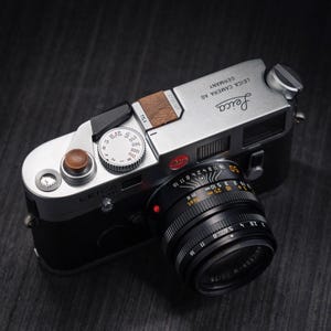 May include: A silver and black Leica M6 film camera with a black lens. The top plate features the Leica logo, the text Leica Camera AG Germany, a wooden shutter release button, a silver shutter speed dial, and a small wooden hot shoe cover.