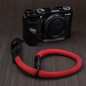 Pode incluir: Uma câmara mirrorless Fujifilm X-M5 preta com tampa de lente, acompanhada por uma correia de pulso trançada em vermelho e preto. A correia apresenta conectores de couro preto e uma pequena peça de destaque vermelha perto do ponto de fixação.