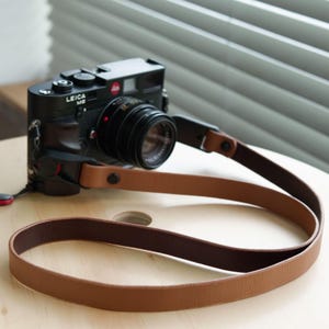 Puede incluir: Una cámara Leica M6 negra con objetivo, que incluye una correa de cuero de dos tonos. La correa es de color tostado en el exterior y marrón oscuro en el interior, sujeta al cuerpo de la cámara con herrajes negros y cierres metálicos redondos.