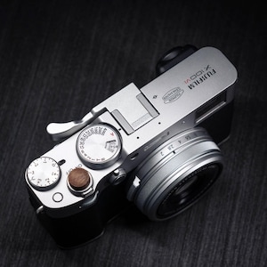 May include: A silver and black Fujifilm X100VI digital camera featuring a top-mounted shutter speed dial, an exposure compensation dial, and a circular wooden shutter release button. The lens barrel displays aperture markings and the Fujinon lens system logo.