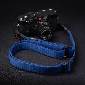 Puede incluir: Una cámara de película Leica M6 negra con objetivo, que cuenta con una correa de tela tejida de color azul brillante. La correa incluye hebillas de ajuste de metal negro y pestañas de cuero con pequeños detalles rojos en los puntos de unión.