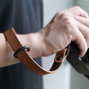 Könnte beinhalten: Ein beigefarbenes Kamerahandgelenkband aus Leder mit glatter Oberfläche, einem verschiebbaren Einstellring aus schwarzem Metall und einem schwarzen Metallnietverschluss, der das Band am Kameragehäuse befestigt und sicheren Halt bietet.