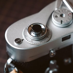 May include: A silver top plate of a vintage film camera featuring a black shutter release button, a circular film advance dial with numerical markings from 0 to 35, and a secondary silver dial with various shutter speed settings and a central hole.