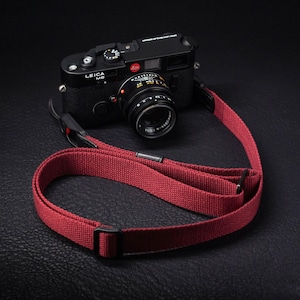 Op de afbeelding: Een zwarte Leica M6 meetzoekercamera met gemonteerde lens, voorzien van de tekst Leica M6 en Ernst Leitz Wetzlar Germany. Aan de camera is een lange, geweven, donkerrode camerariem bevestigd met zwarte metalen verstelgespen en verbindingsstukken.