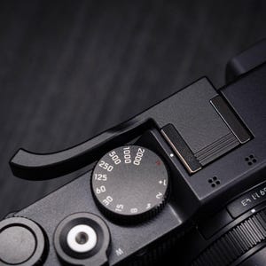Pode incluir: O topo de uma câmara digital em preto mate com um seletor circular de velocidade do obturador com marcações numéricas brancas de 8 a 2000, uma sapata de flash, um apoio para o polegar curvo e um botão de disparo com centro prateado.