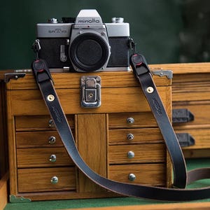 Op de afbeelding: Een zilveren en zwarte Minolta SRT 101 filmcamera met een zwarte lensdop, bevestigd aan een zwarte leren camerariem met goudkleurige metalen klinknagels en rode accenten op de verstelbare clips, rustend op een houten opbergkist.