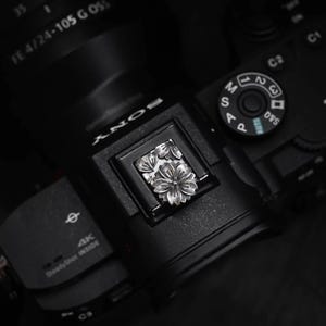 Peut inclure: Un cache-griffe en métal argenté orné d'un motif floral détaillé avec des pétales en relief, inséré dans la griffe porte-accessoire d'un boîtier d'appareil photo numérique noir avec un objectif et divers cadrans de contrôle visibles.