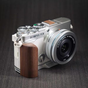 May include: An Olympus Pen digital camera featuring a silver and white body with a dark brown wooden grip. The camera is equipped with an M.Zuiko Digital 14-42mm lens. The top panel includes silver dials, a shutter button, and a rectangular accessory port.