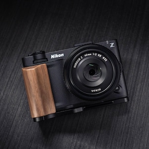 Peut inclure: Un appareil photo hybride Nikon Z noir doté d'une poignée texturée et d'une extension en bois. L'objectif est un Nikkor Z 40mm 1:2 SE avec un diamètre de filtre de 52mm. Le boîtier affiche le logo Nikon et l'emblème de la série Z en haut à droite.