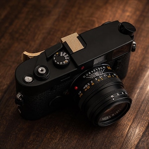 Pode incluir: Uma câmara telémetrica preta com uma cobertura de sapata de cor dourada texturizada e uma alavanca de avanço de filme dourada a condizer. A câmara está equipada com uma lente preta com marcações numéricas brancas e cor de laranja.