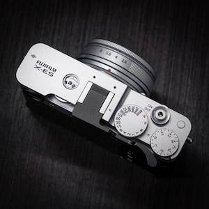 Peut inclure: Un appareil photo numérique Fujifilm X-E5 argenté vu du dessus, doté d'un objectif avec des ouvertures visibles, d'une molette de vitesse d'obturation de 15 à 4000, d'une molette de correction d'exposition et d'un bouton d'alimentation intégré.