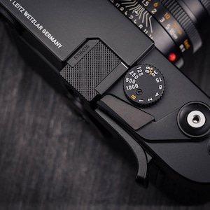 Puede incluir: Un cuerpo de cámara Leica negro con el texto LEITZ WETZLAR GERMANY, el número de serie 5706036 y una tapa de zapata de metal texturizado. La placa superior incluye un dial de velocidad de obturación con números blancos y una B y rayo amarillos.