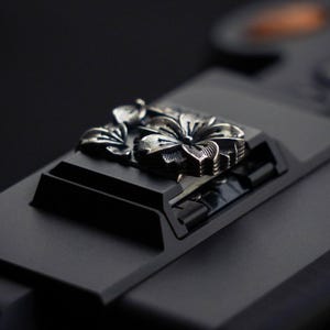 May include: A metallic silver-toned floral ornament featuring detailed petal engravings, securely attached to a matte black rectangular base. The floral design displays a central bloom with surrounding leaves, showcasing a high-contrast finish.