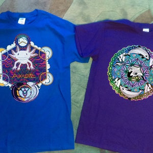 May include: Two t-shirts, one blue and one purple. The blue shirt features a colorful axolotl design with the word "AXOLOTL". The purple shirt has a circular design with stylized dolphins.