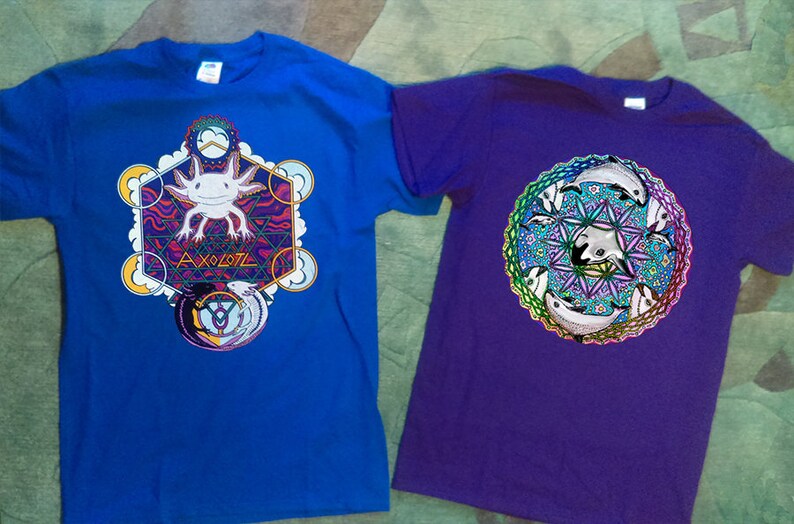 May include: Two t-shirts, one blue and one purple. The blue shirt features a colorful axolotl design with the word "AXOLOTL". The purple shirt has a circular design with stylized dolphins.