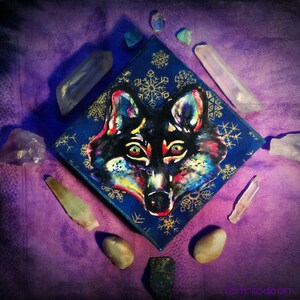 Könnte beinhalten: Ein farbenfrohes Gemälde eines Wolfskopfes auf einer blauen Leinwand mit goldenen Schneeflockenmustern. Das Gemälde ist von Kristallen auf einem violetten Hintergrund umgeben.