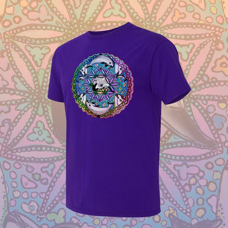 May include: A purple t-shirt featuring a circular design with white and black orcas, geometric shapes, and a rainbow border. The orcas are arranged around a central star pattern. The shirt has short sleeves and a crew neck.