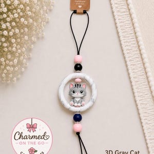 May include: A hanging car charm featuring a gray 3D cat figurine centered within a white marble-patterned ring. The charm is suspended by a black cord with pink, black, and navy blue beads, and includes a brown tag labeled Handmade with love.
