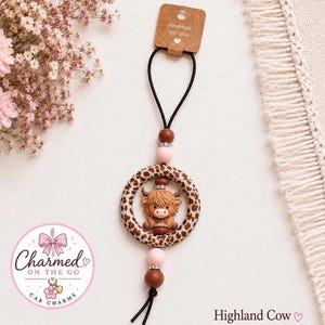 May include: A car charm featuring a brown Highland cow figurine centered within a leopard print ring. The charm includes brown and light pink beads with silver accents, suspended by a black cord attached to a brown tag labeled Handmade with love.