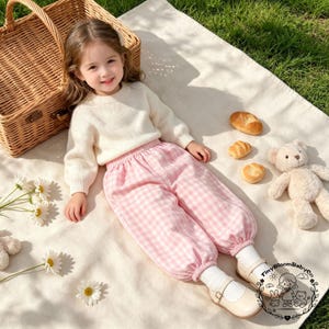 May include: A long-sleeved cream-colored knit sweater paired with pink and white gingham pants featuring an elastic waistband and cuffed ankles. The outfit is completed with white socks and beige buckled shoes, showcasing a soft, textured aesthetic.