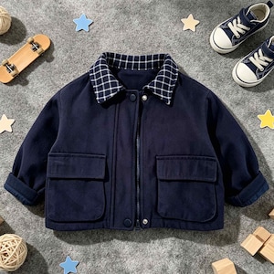 Peut inclure: Une veste bleu marine foncé dotée d'un col rabattu à motif quadrillé blanc, d'une fermeture éclair centrale, de deux grandes poches plaquées à l'avant et de manches longues à revers. La veste est confectionnée dans un tissu uni et lisse.