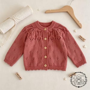 May include: A dusty rose long-sleeve knit cardigan featuring a round neckline, five light-colored buttons, and a delicate openwork pattern across the chest and sleeves. The garment has ribbed cuffs and a ribbed hem with a scalloped edge finish.