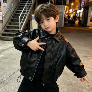 May include: A black faux leather jacket featuring a classic collar, long sleeves with buttoned cuffs, a front zip fastening, and vertical seam detailing. The jacket has a structured fit with a smooth finish and a black interior lining visible at the collar.