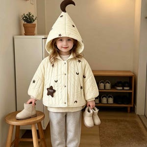 Puede incluir: Una chaqueta acolchada de color crema con una capucha puntiaguda rematada con una luna creciente de color marrón oscuro. La chaqueta está decorada con pequeñas estrellas y lunas bordadas, un parche de estrella grande y botones a presión.
