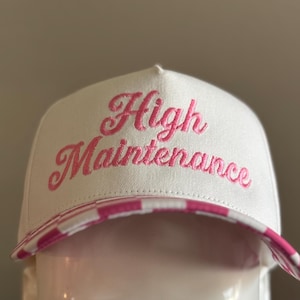 May include: A white baseball cap featuring the embroidered pink cursive text High Maintenance on the front panel. The brim of the cap displays a pink and white checkered pattern. The hat has a structured crown and a curved visor.