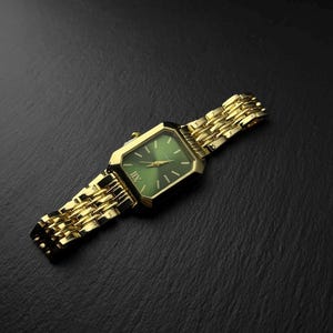 Pode incluir: Um relógio de pulso dourado com uma caixa retangular e um mostrador verde profundo. O relógio possui uma pulseira de elos metálicos com acabamento polido. O mostrador exibe marcadores de hora e ponteiros dourados, com um algarismo romano XII.