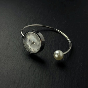 May include: A silver-toned cuff bracelet featuring a round watch face with a white dial and silver hands on one end, and a single white spherical pearl accent on the opposite open end. The watch face has a faceted glass cover and silver markers.