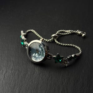 Peut inclure: Une montre à quartz argentée avec un cadran rond en nacre. Le bracelet à chaîne réglable est orné d'étoiles décoratives argentées, agrémentées de cristaux verts et de strass transparents, formant un motif d'inspiration céleste.