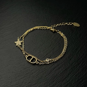 May include: A gold-tone double chain bracelet featuring a central CD logo charm, a small star charm encrusted with clear stones, and a single round clear stone accent. The bracelet includes a delicate link chain and a small oval tag at the clasp.