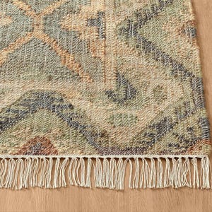 May include: A woven area rug featuring a geometric pattern in muted shades of sage green, dusty blue, and soft peach on a cream background. The rug has a textured, knotted surface and is finished with a row of long, cream-colored fringe tassels.