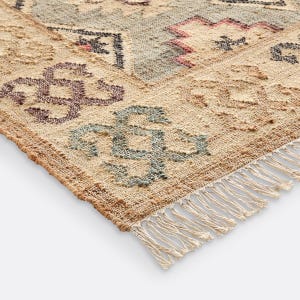Puede incluir: Una alfombra tejida con una base beige texturizada y patrones geométricos en tonos azul apagado, rojo oscuro y gris carbón. La alfombra presenta un tejido anudado visible y un borde con flecos en la parte inferior, mostrando un diseño detallado.
