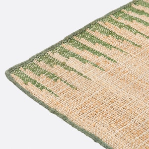 May include: A woven rug featuring a textured surface with a natural tan base color and vertical stripes in a muted green tone. The rug has a finished edge with a matching green fabric border that runs along the perimeter of the rectangular textile.