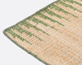 May include: A woven rug featuring a textured surface with a natural tan base color and vertical stripes in a muted green tone. The rug has a finished edge with a matching green fabric border that runs along the perimeter of the rectangular textile.