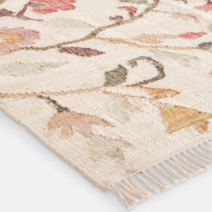May include: A woven area rug featuring a cream-colored base with a repeating floral pattern in muted shades of pink, orange, and sage green. The rug has a textured surface and is finished with short, cream-colored fringe along the bottom edge.