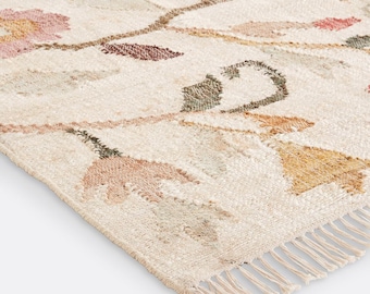 May include: A woven area rug featuring a cream-colored base with a repeating floral pattern in muted shades of pink, orange, and sage green. The rug has a textured surface and is finished with short, cream-colored fringe along the bottom edge.